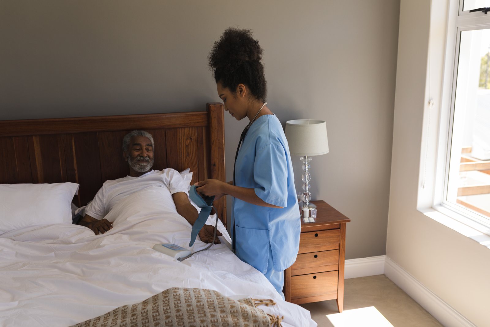 A nurse attends to an older man resting in bed at home during a home visit